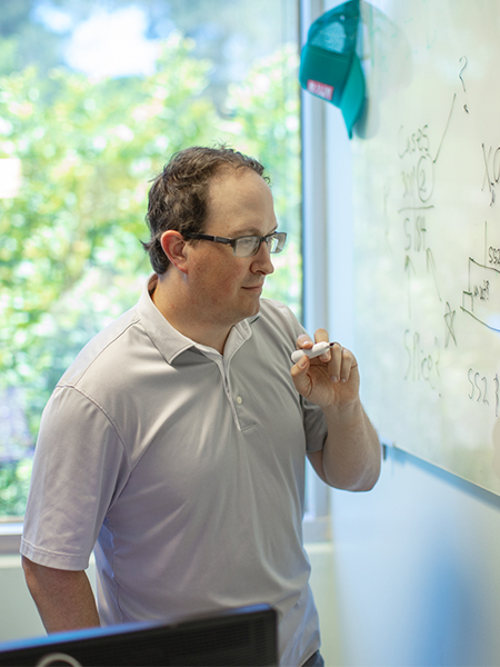 A person writing on a whiteboard on the wall of their office with an Ambry hat hanging in the background